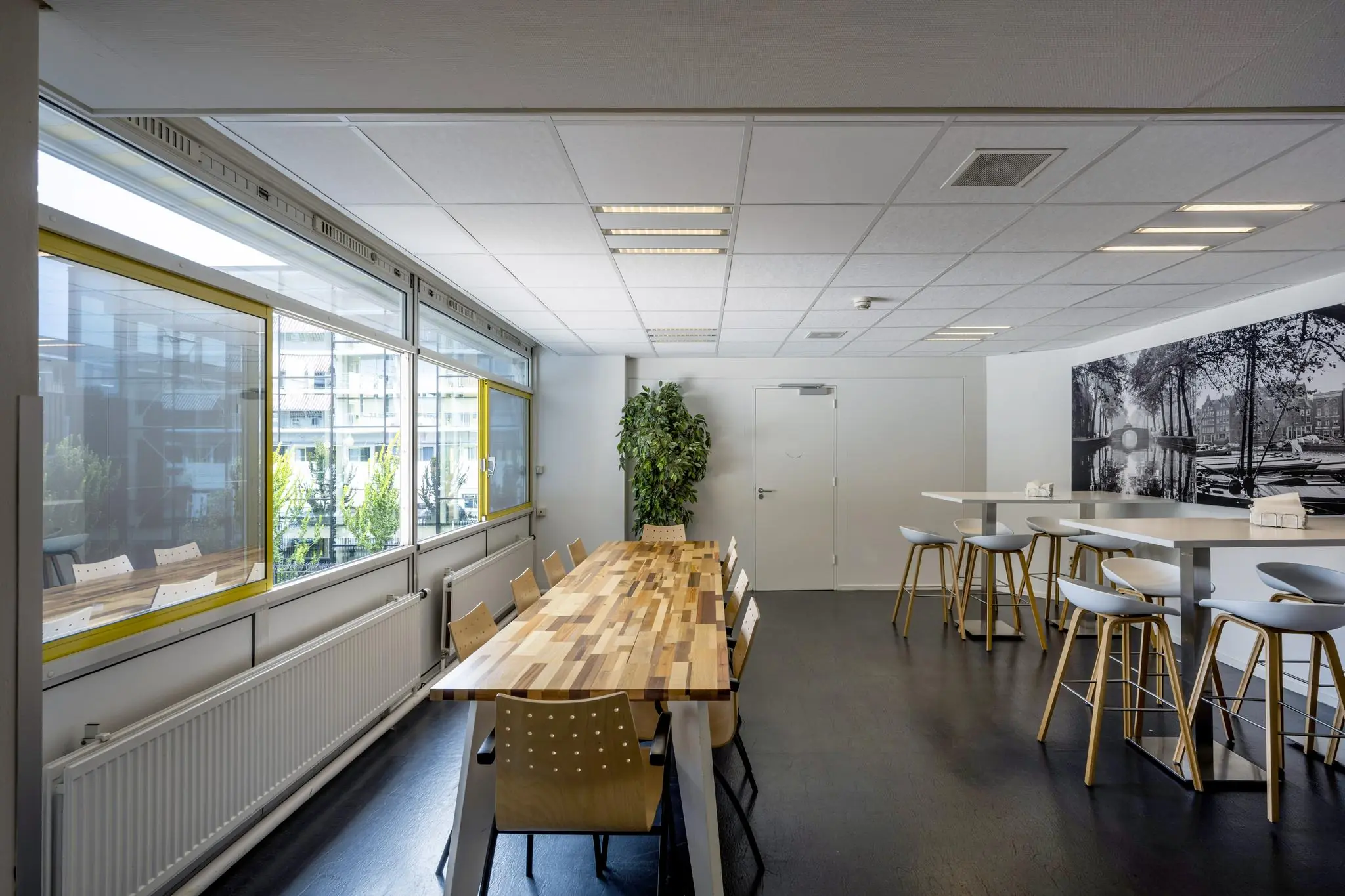 Moderne kantineruimte aan de Jan Tooropstraat met grote ramen, houten tafel, barkrukken en een zwart-wit muurfoto van een Amsterdamse gracht.